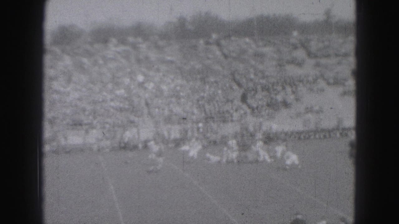 Distant black-and-white 8mm view of a Harrisburg football game, with players spread across the field and a large crowd in the stands.