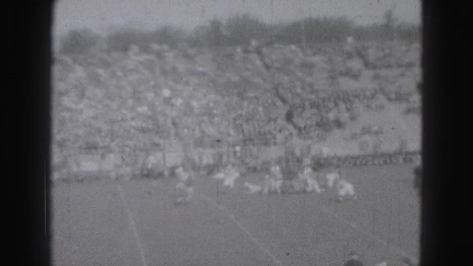 Distant black-and-white 8mm view of a Harrisburg football game, with players spread across the field and a large crowd in the stands.