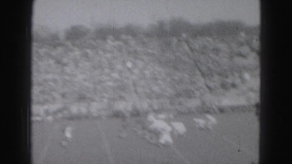 Black-and-white 8mm frame from a 1975 Harrisburg football game, showing blurred players near the lower field and a dense spectator section beyond.
