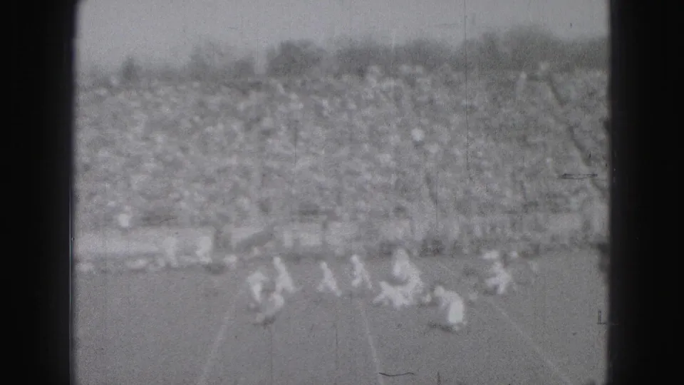 High-angle 1975 Harrisburg football view with players clustered near the lower edge, bleachers in the background, and visible black-and-white film wear.