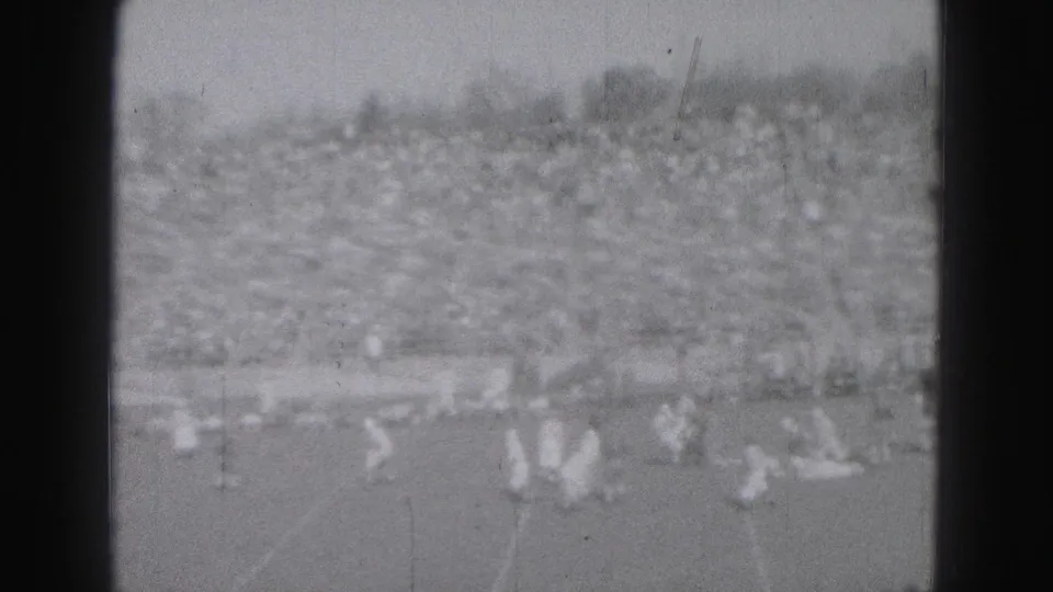 1975 Harrisburg football game scene with players spread across the field and a packed stand in soft black-and-white 8mm footage.