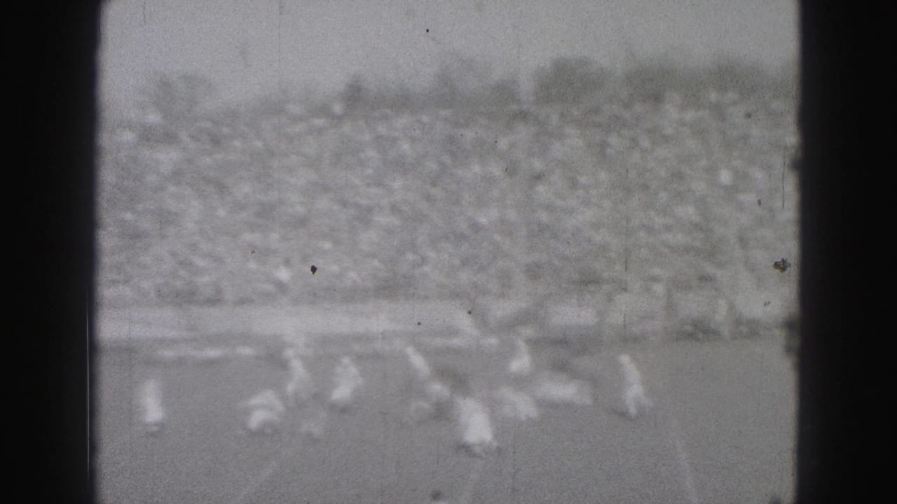 Players move through a pass play on a Harrisburg football field, with a dense spectator crowd behind them in 1975 8mm footage.