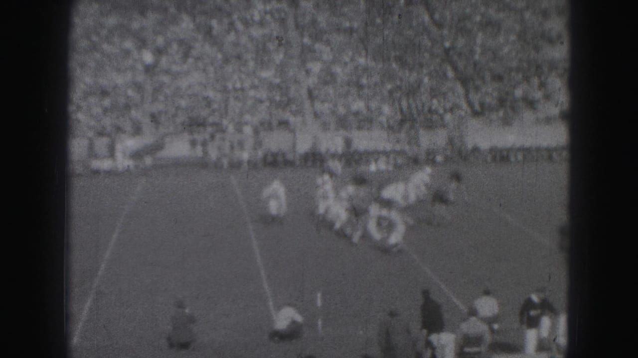 Football players collide near a line of scrimmage with spectators in the background.