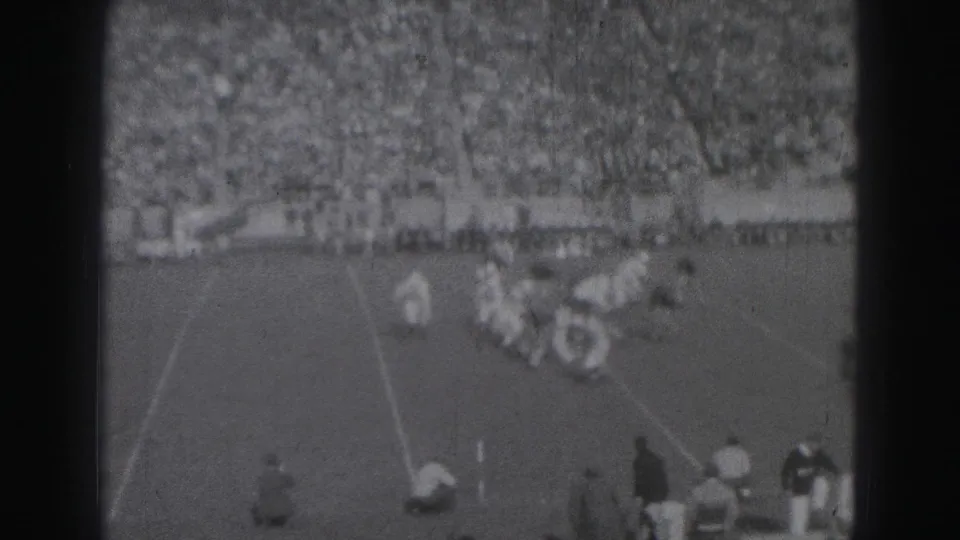 1975 Harrisburg 8mm footage shows football players in light and dark uniforms meeting near the line of scrimmage before crowded bleachers.