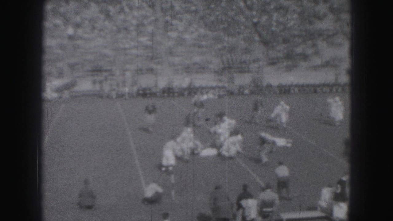 Football players gather around a tackle or blocking play on a grass field, with Harrisburg spectators visible in the distant stands.