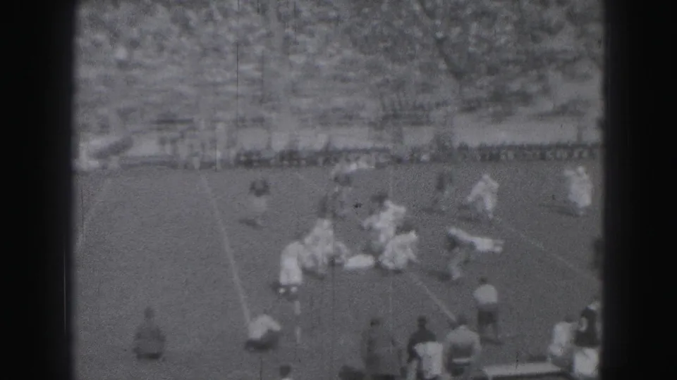 Football players gather around a tackle or blocking play on a grass field, with Harrisburg spectators visible in the distant stands.