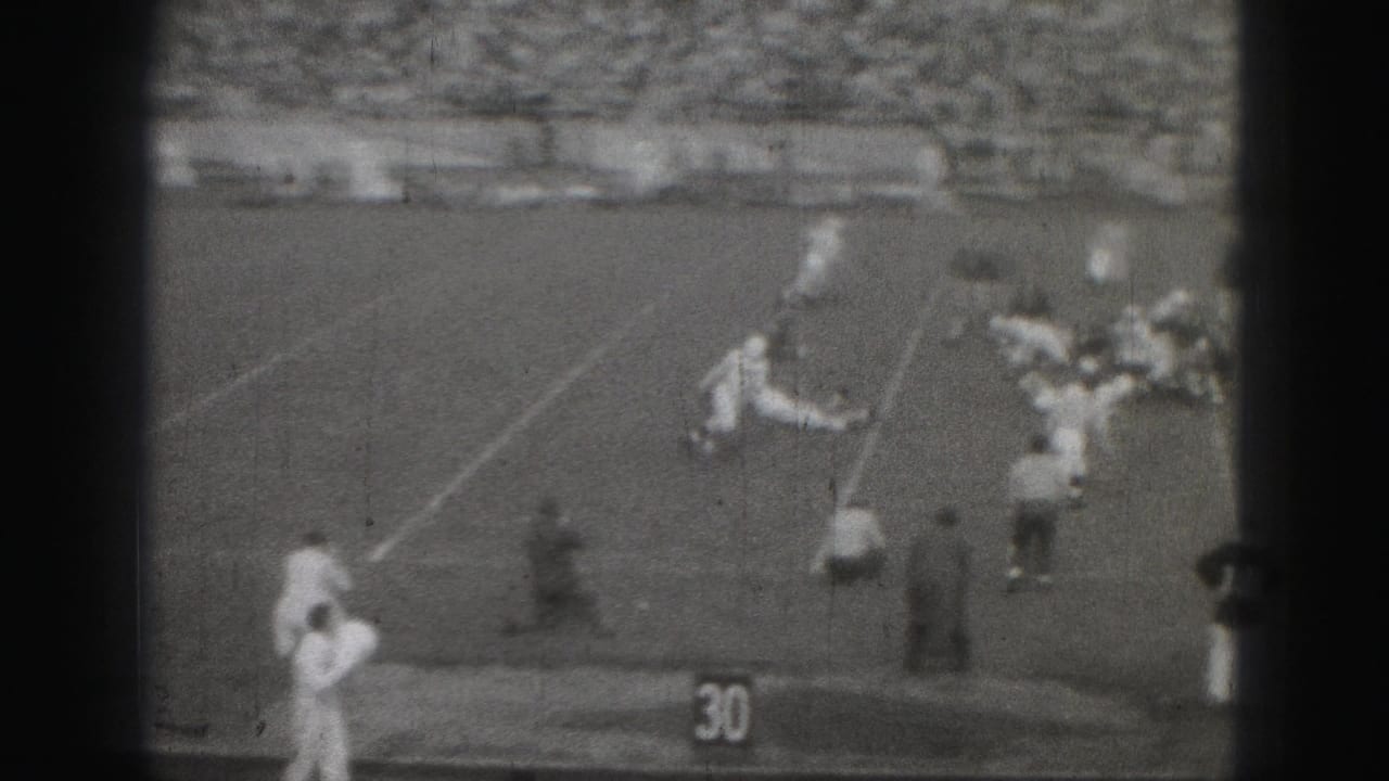 Distant 1975 Harrisburg football action near the sideline, with players in light and dark uniforms and a packed grandstand.