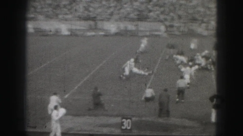 Distant 1975 Harrisburg football action near the sideline, with players in light and dark uniforms and a packed grandstand.