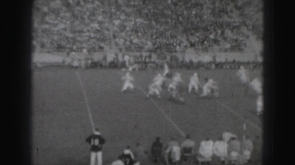 Distant 1975 Harrisburg football action shows players converging near the sideline as the crowd watches from the stands.