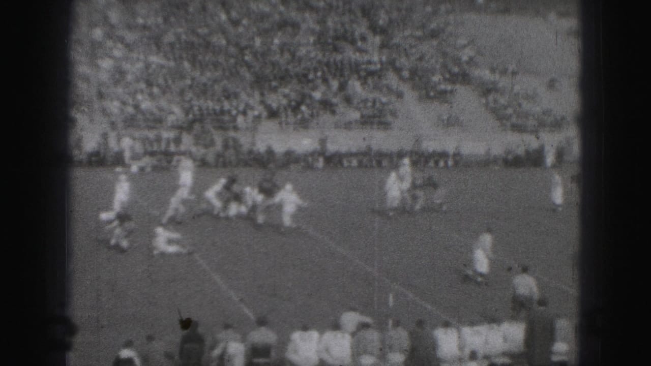 Wide football field view with players in white uniforms and spectators in bleachers.