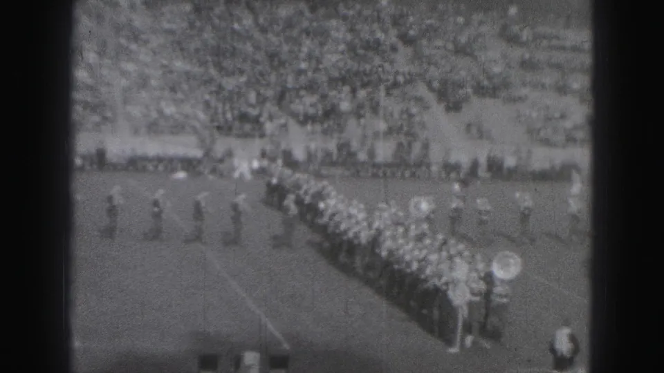 Distant 1975 Harrisburg frame showing marching band rows and spectators during a school football event.