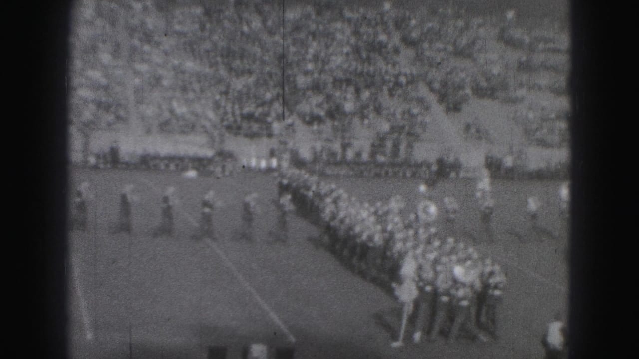 Marching band lines up on a football field with spectators in the stands.