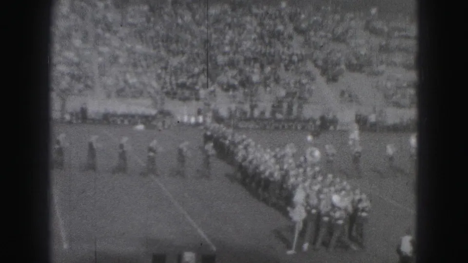 Distant 1975 Harrisburg football-field view with a marching band in formation and a crowded grandstand behind it.
