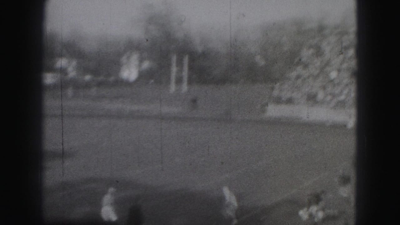 Soft black-and-white view of a football field with players near the lower edge and a goalpost beyond.