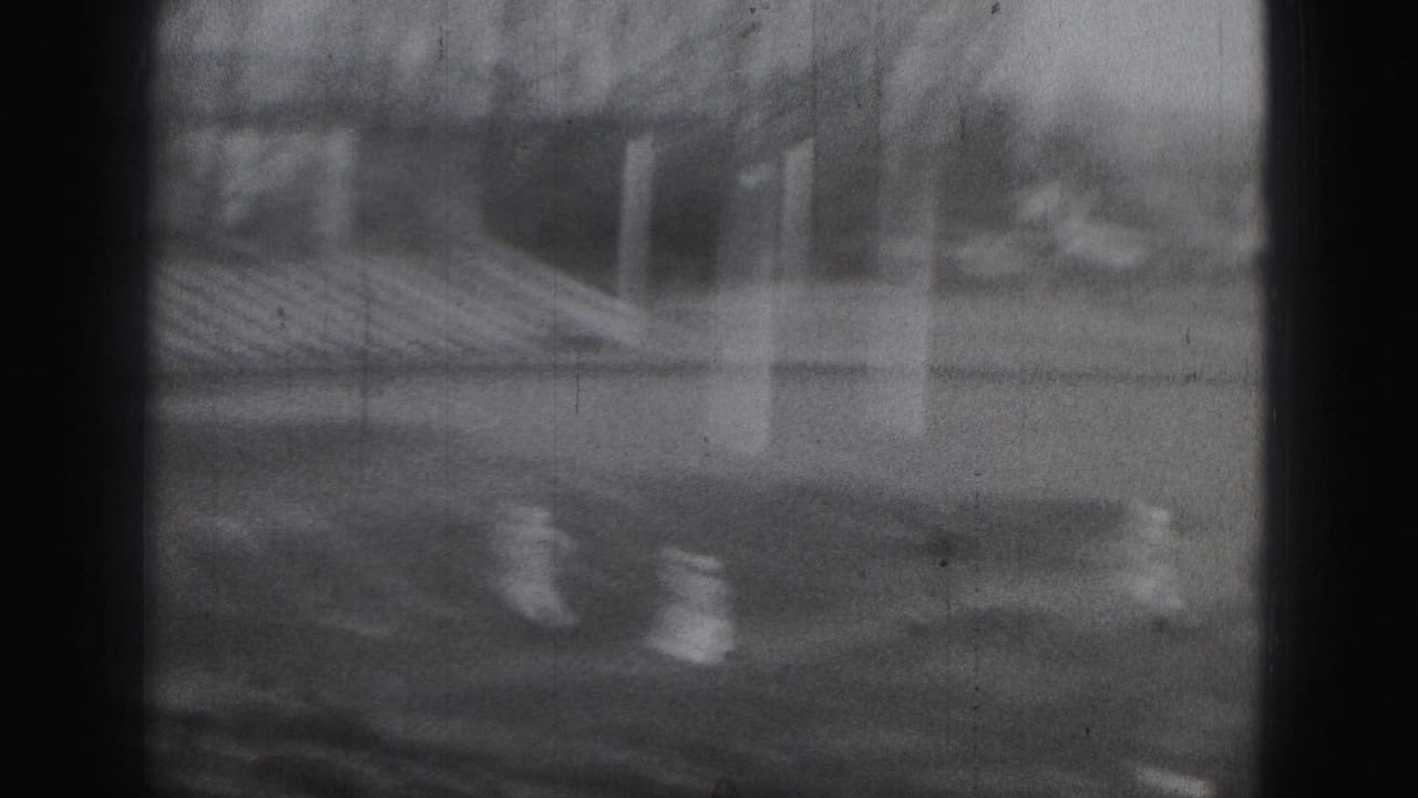 Black-and-white 8mm frame from the Harrisburg football game, showing players near the goalpost and stadium seating beyond.