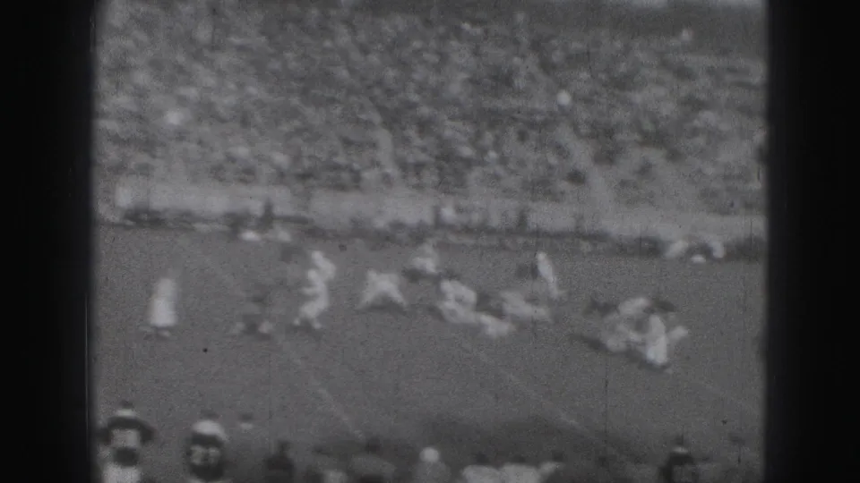 Black-and-white 8mm frame from the Harrisburg game showing players breaking from the line with bleachers in the background.