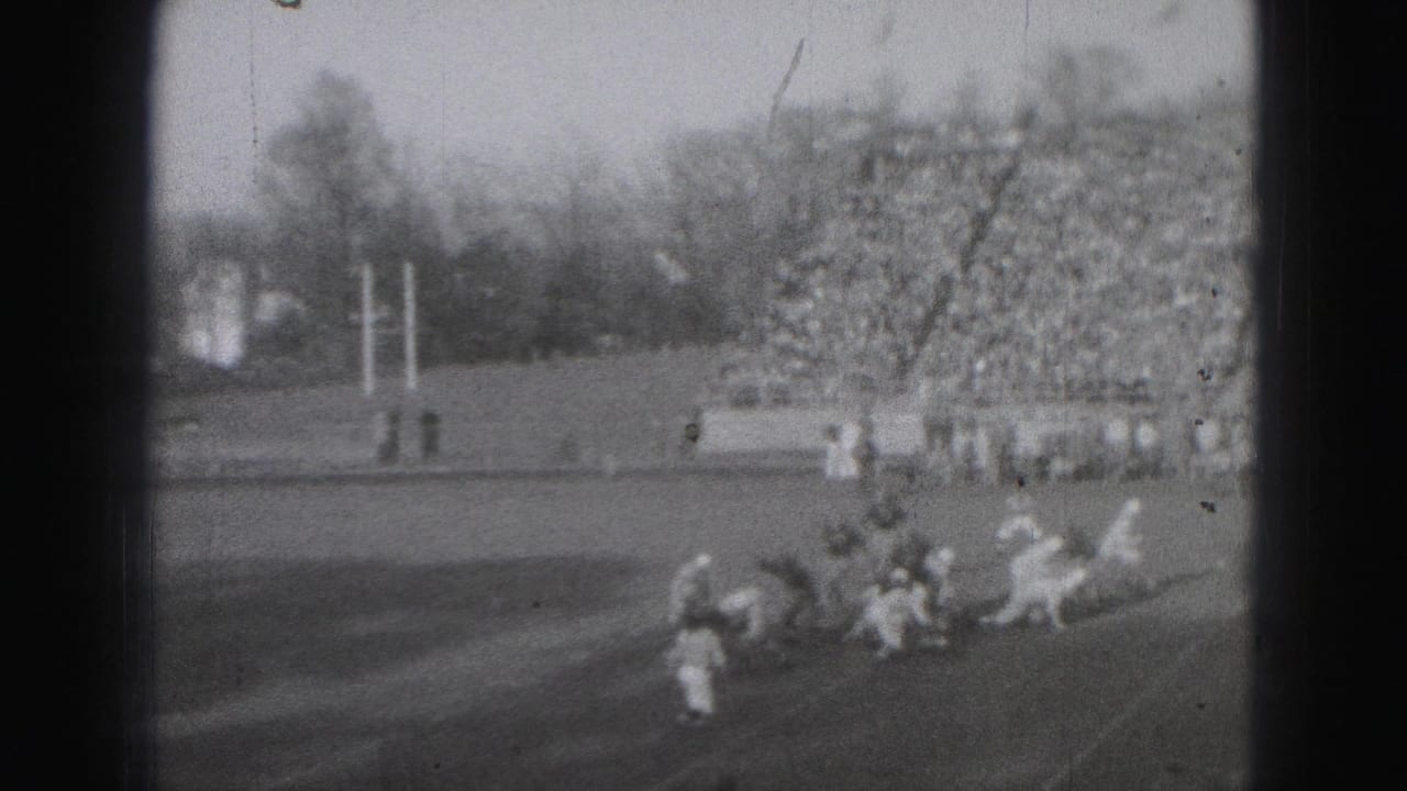 1975 Harrisburg football game footage showing players in light and dark uniforms clustered near the line as spectators fill the stands.
