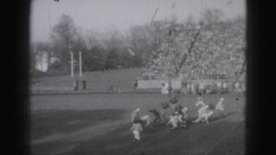 1975 Harrisburg football game footage showing players in light and dark uniforms clustered near the line as spectators fill the stands.