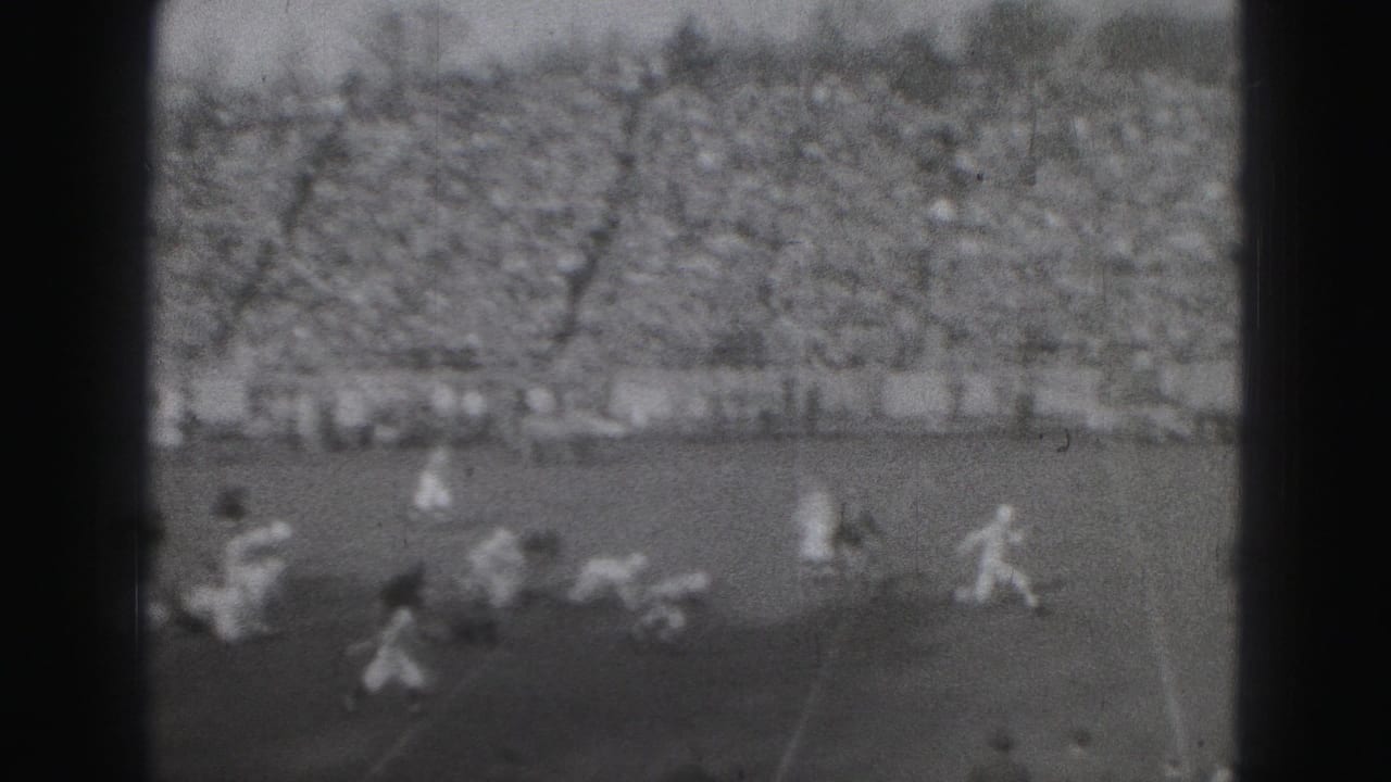 Wide 8mm view of a 1975 Harrisburg football play with players spread across the field and a dense stadium crowd behind them.