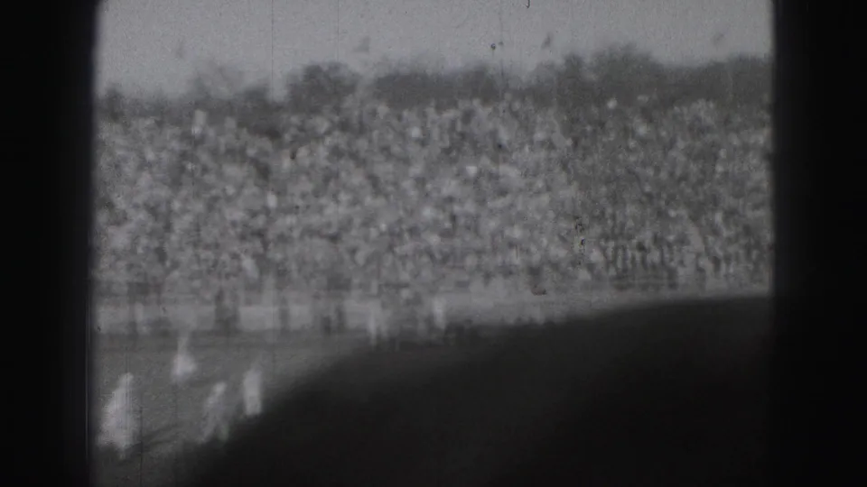 Grainy black-and-white 8mm frame from the Harrisburg football game, with players low in frame and spectators filling the stands.