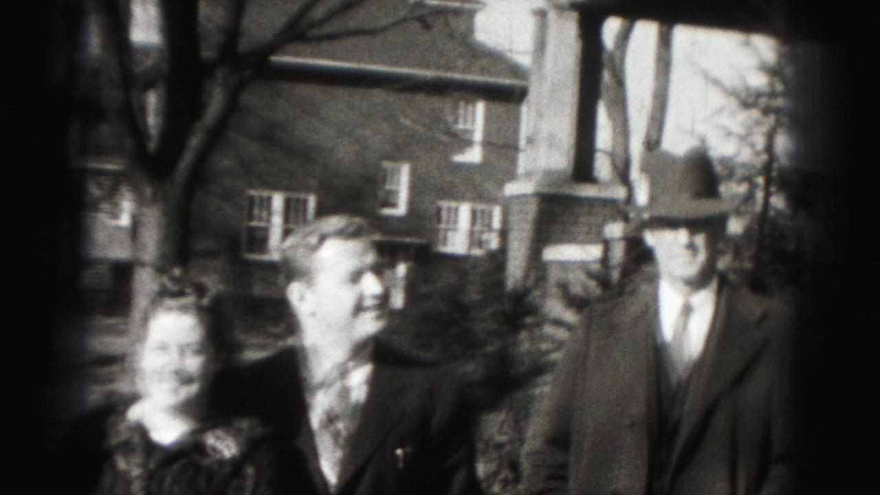 In 1975 Harrisburg home-movie footage, three adults stand near a brick house while looking toward the camera.