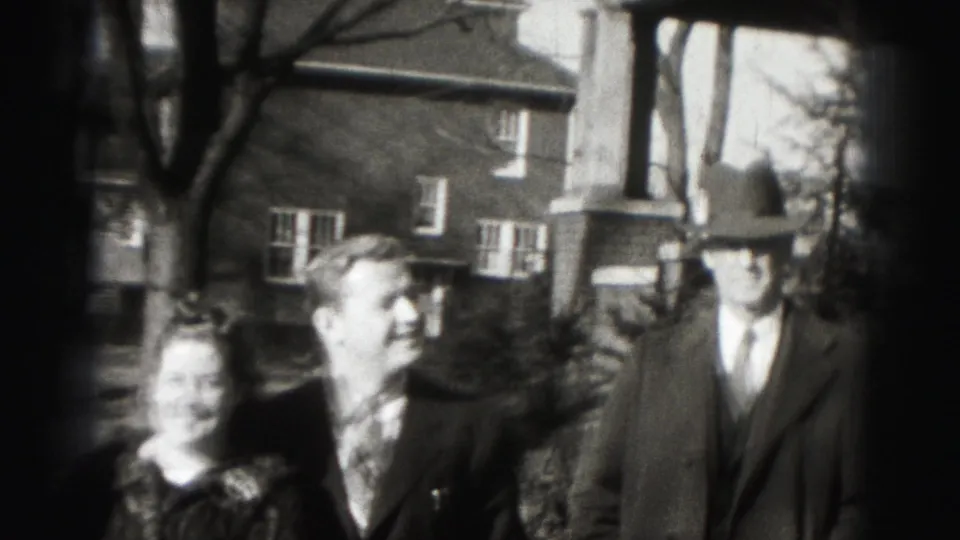 In 1975 Harrisburg home-movie footage, three adults stand near a brick house while looking toward the camera.