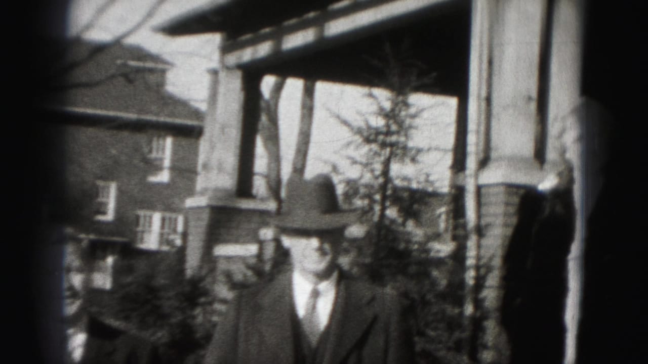 A man in a fedora and suit stands near porch columns beside a brick house.