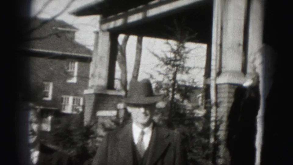 A formally dressed man in a fedora stands near the porch of a Harrisburg brick house in 1975 home-movie footage.