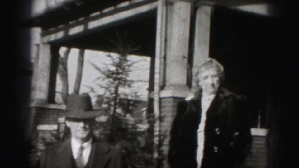 The camera frames a man in a fedora and a woman in a dark coat outside the brick house in Harrisburg.