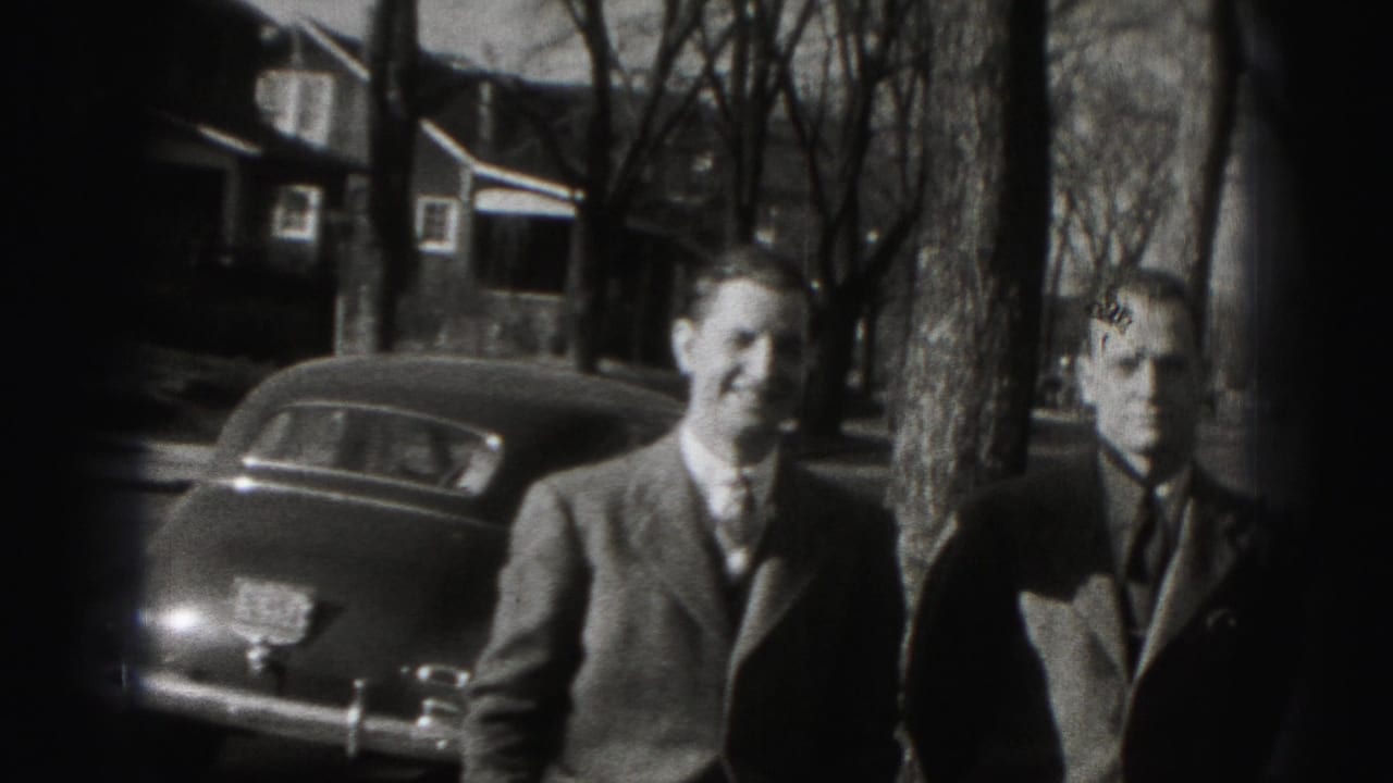 In Harrisburg home-movie footage, three suited adult men stand by a parked car with houses and bare trees behind them.