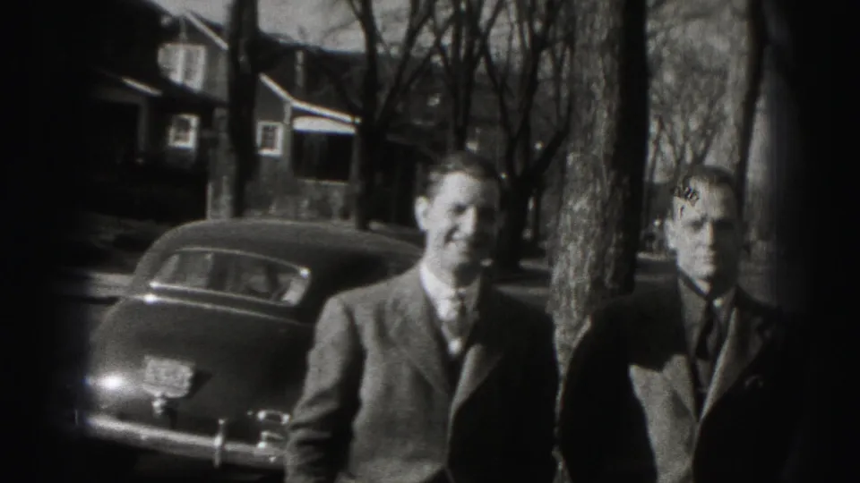 In Harrisburg home-movie footage, three suited adult men stand by a parked car with houses and bare trees behind them.