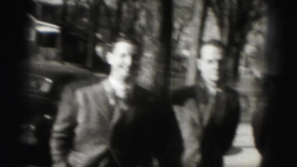 Two suited adult men are seen in soft focus on a Harrisburg sidewalk, with trees and a parked car in the background.