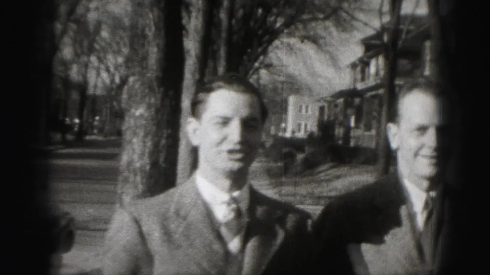 Two suited adult men stand on a tree-lined Harrisburg sidewalk, with houses and a street receding behind them.