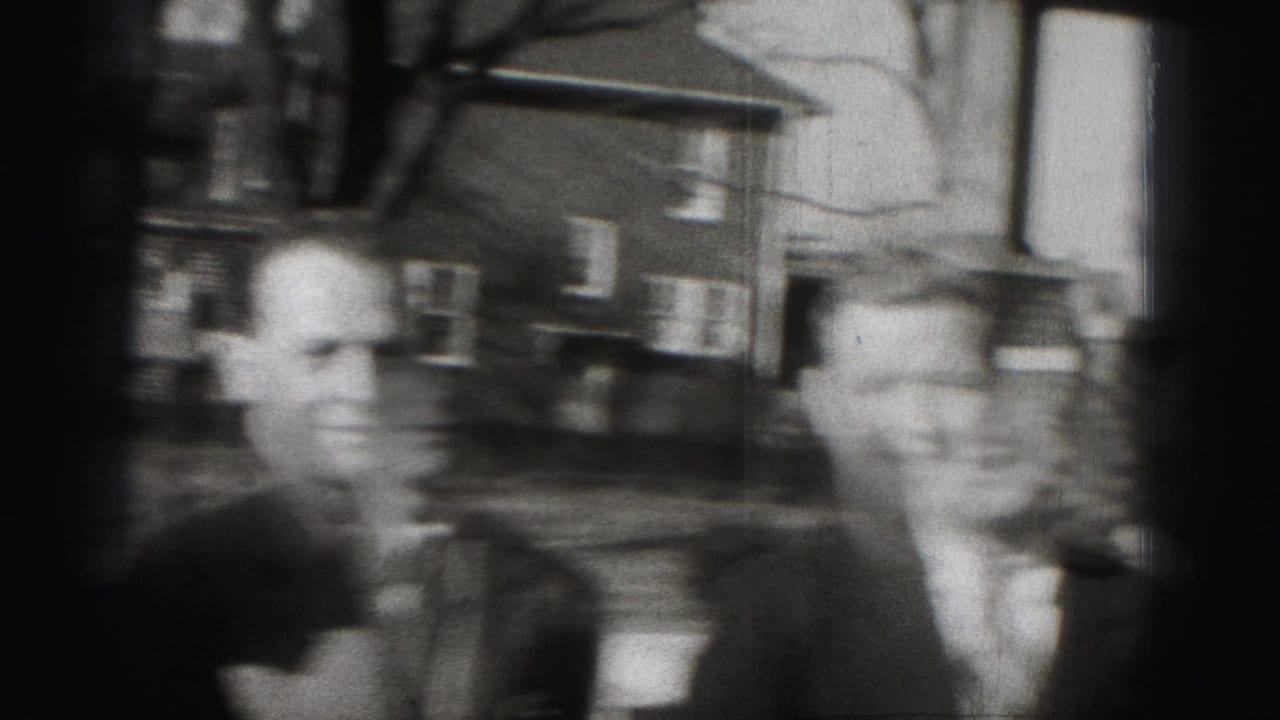 Two suited men standing outdoors near houses and bare trees.