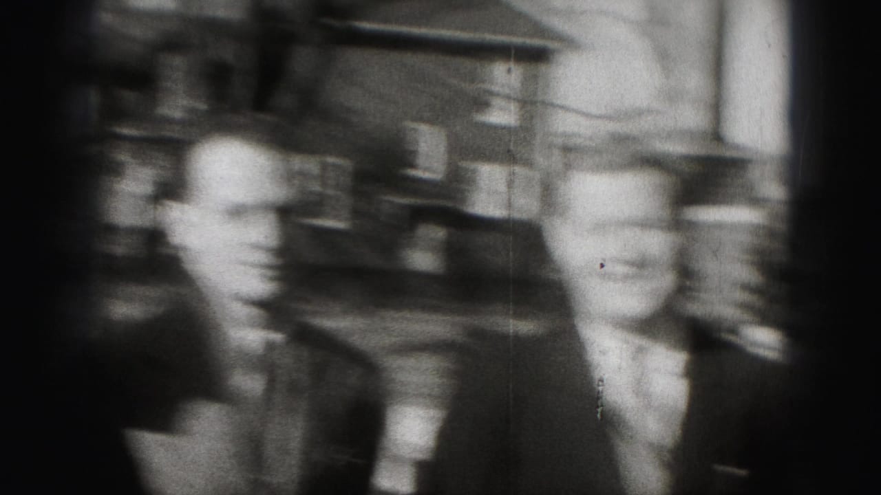 Two suited men stand close together outdoors with houses behind them.