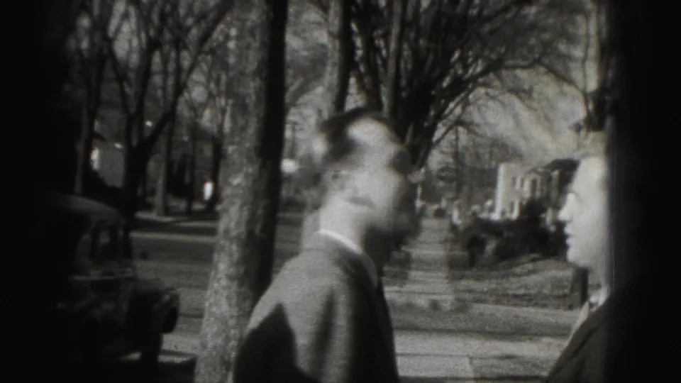A suited man turns near a tree-lined residential sidewalk in the Harrisburg home-movie clip.