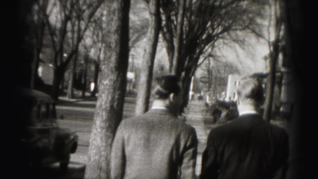 The two men continue along the sidewalk, seen from behind with vintage cars and bare street trees nearby.