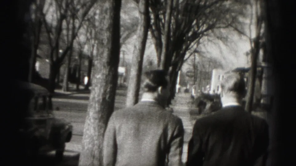 The two men continue along the sidewalk, seen from behind with vintage cars and bare street trees nearby.