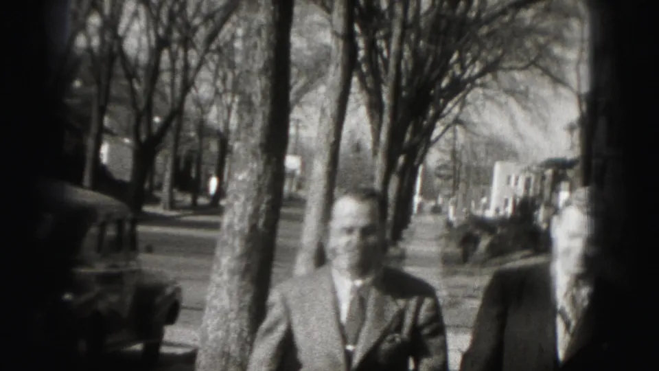 The men face the camera at the end of the short sidewalk shot, with leafless trees and a parked car behind them.