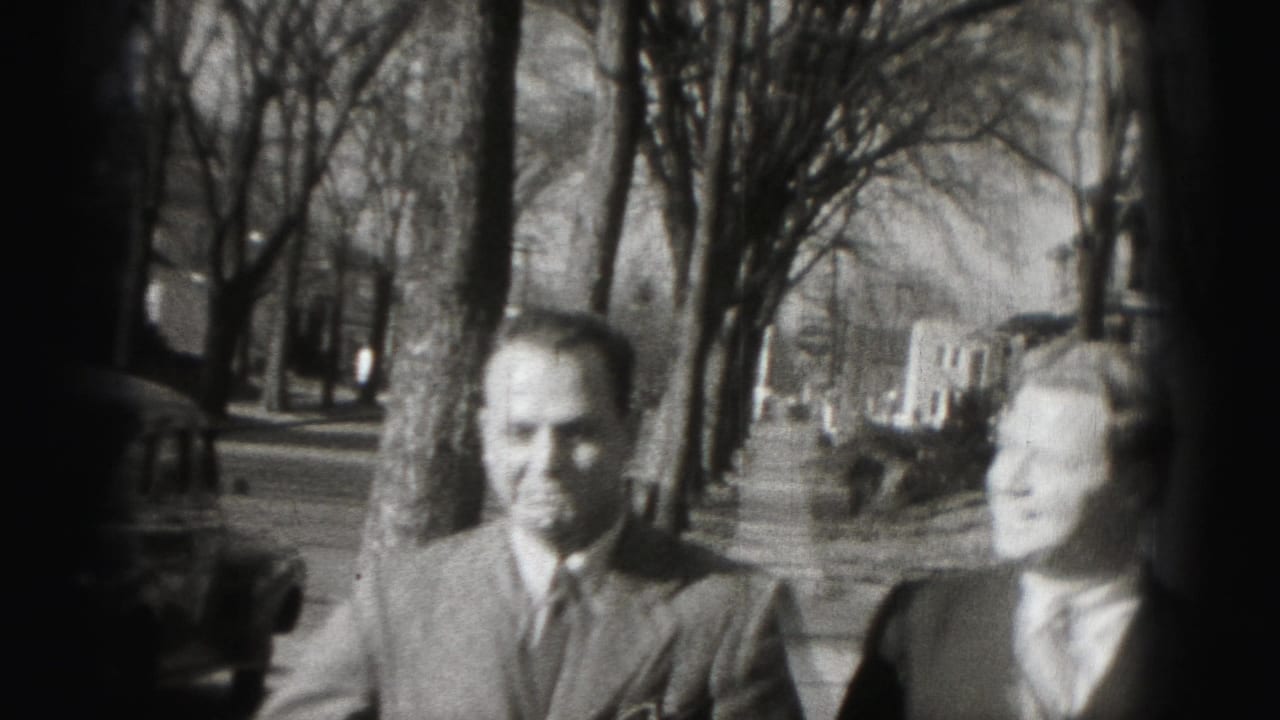 Two suited men walk along a tree-lined sidewalk with parked cars and houses nearby.