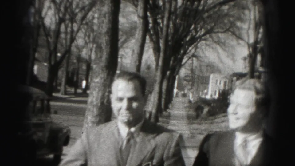 In 1975 Harrisburg footage, two adult men in suits walk on a residential sidewalk beside bare trees and parked cars.