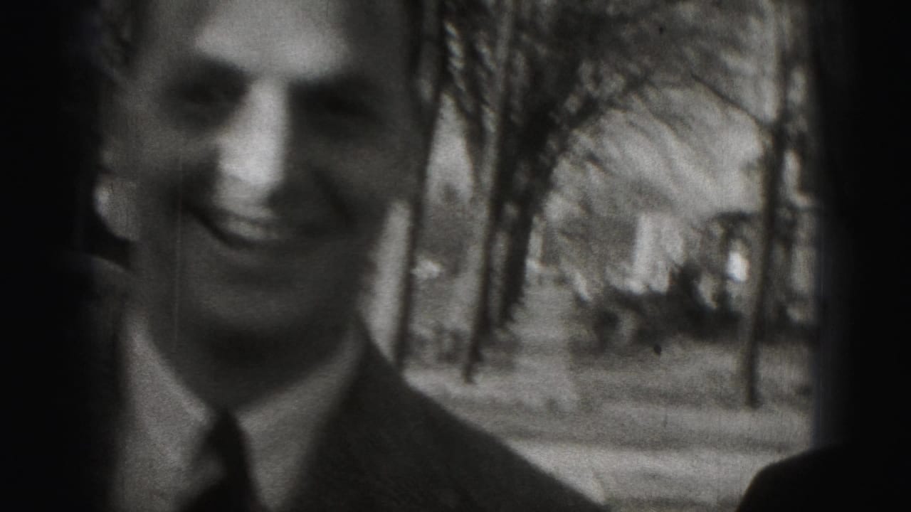 A smiling suited man appears close to the camera on a sidewalk framed by trees.
