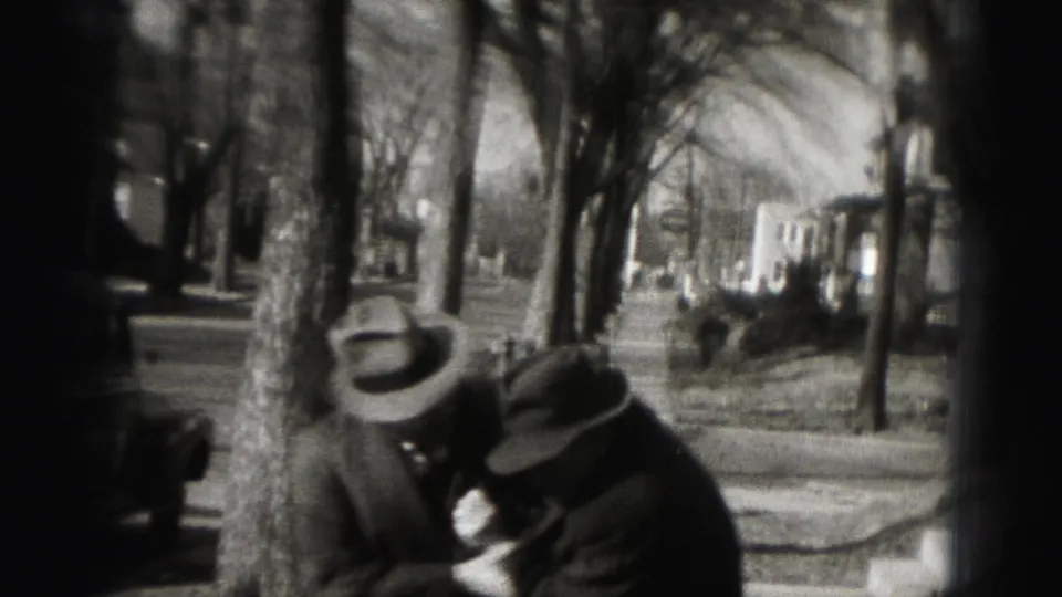 Two adult men wearing brimmed hats lean near the camera on a tree-lined Harrisburg sidewalk in 1975 home-movie footage.