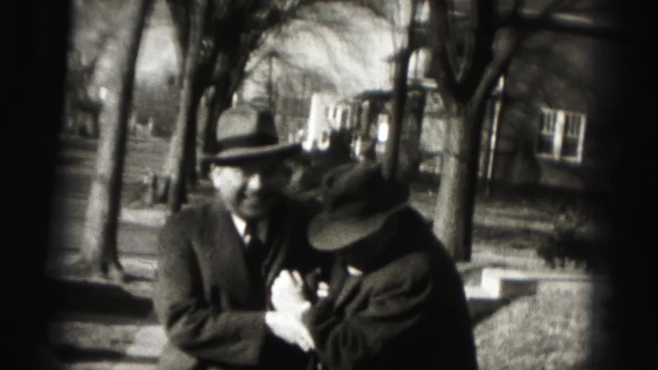 Two adult men in hats and dark coats stand together on a Harrisburg sidewalk, with one adjusting his chest or lapel area.