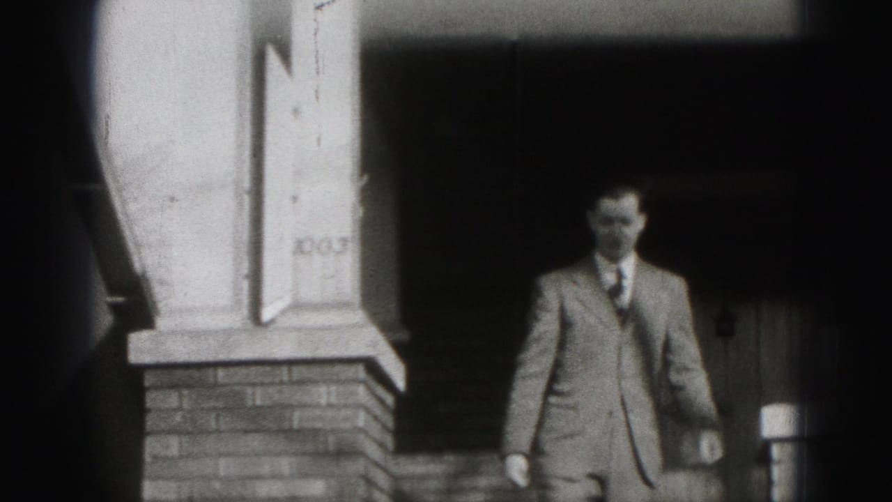 Man in a suit stands near brick porch columns and shaded steps.