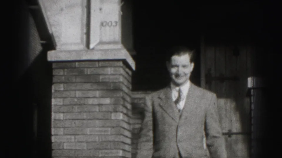 A suited adult man moves down or away from the porch area of a Harrisburg house in this 1975 8mm frame.