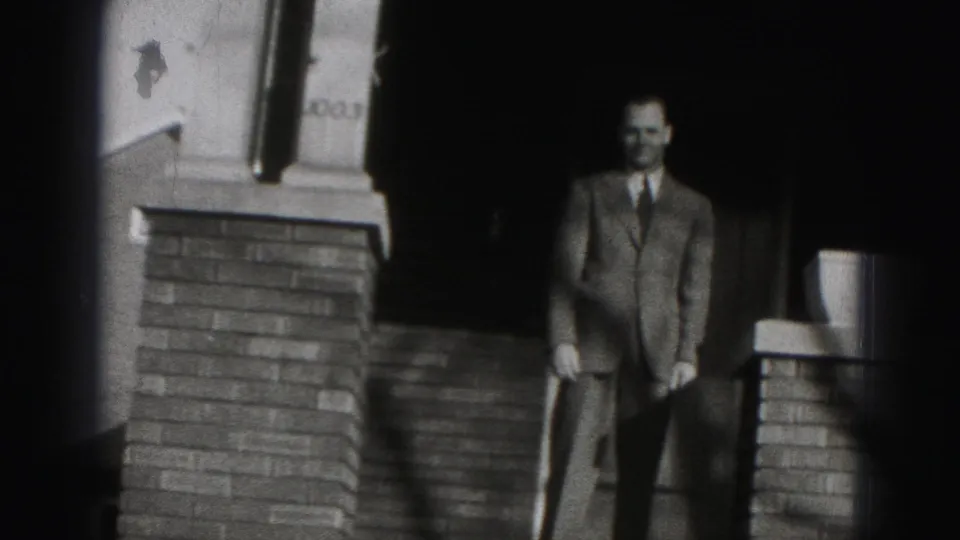 The suited man stands at or near the bottom of the brick porch steps outside a Harrisburg house in 1975 footage.