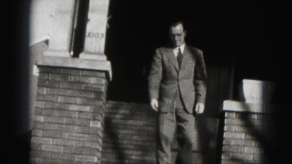 In 1975 Harrisburg home-movie footage, a suited man appears in a doorway framed by brick porch columns.