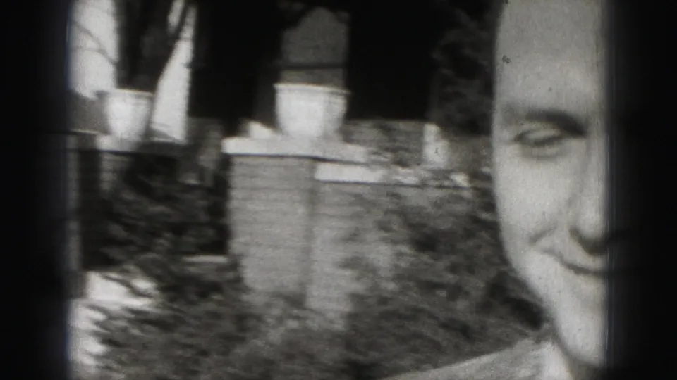Adult man smiles near the 8mm camera in a Harrisburg residential setting, with a tree trunk and house details behind him.