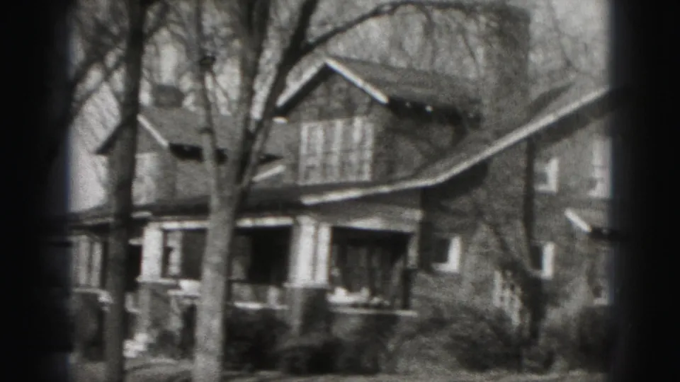 Handheld 8mm view of a Harrisburg residential exterior, showing a porch, columns, bare tree branches and front shrubs.